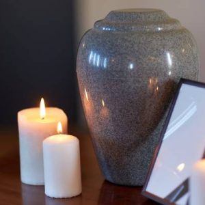 A close-up of a grey cremation urn on a wooden table next to burning white candles and a blank picture frame.