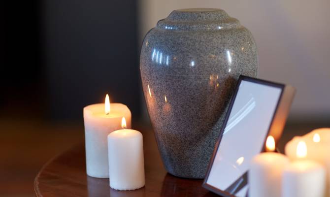 A close-up of a grey cremation urn on a wooden table next to burning white candles and a blank picture frame.
