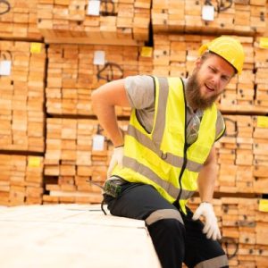 A worker in yellow high-visibility gear is sitting on a bundle of wood planks as he holds his back.