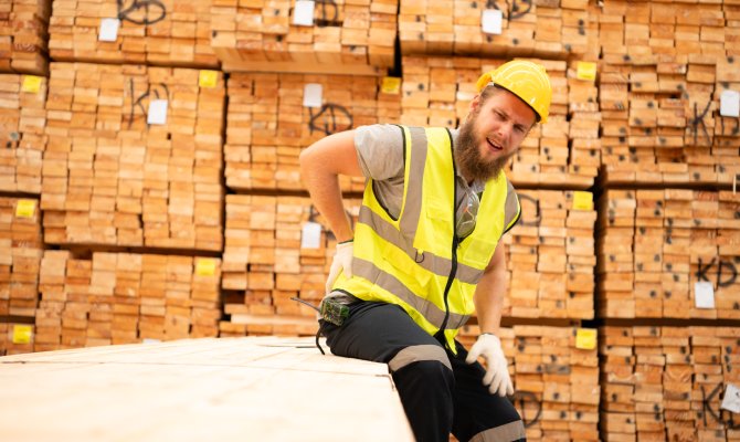 A worker in yellow high-visibility gear is sitting on a bundle of wood planks as he holds his back.