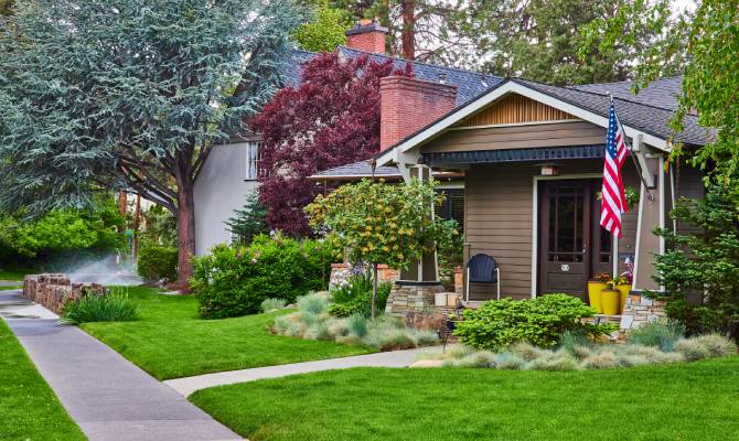 A front yard has thick green grass and ornamental shrubs and trees. An American flag is mounted near the front door.