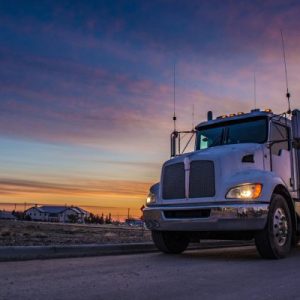 A close-up of a dump truck driving with their headlights on as the sun sets in the background. The sky is pink and blue.
