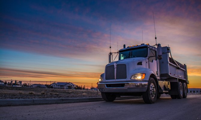 A close-up of a dump truck driving with their headlights on as the sun sets in the background. The sky is pink and blue.