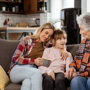 A young girl sits between her mom and grandma as they talk to each other on a couch. Behind them is a kitchen with open shelves.