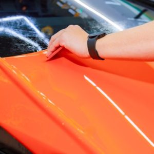 Someone smoothly applying a vinyl wrap to the surface of a vehicle with a shiny orange colored finish.