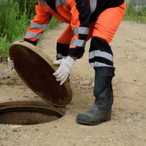 A professional in an orange safety suit takes the cover off a manhole. An orange hard hat sits on the ground.