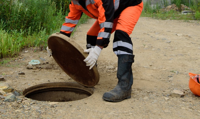 A professional in an orange safety suit takes the cover off a manhole. An orange hard hat sits on the ground.