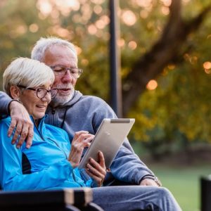 An older man and woman sit together on a park bench and look at a tablet device. A tree stands behind them.