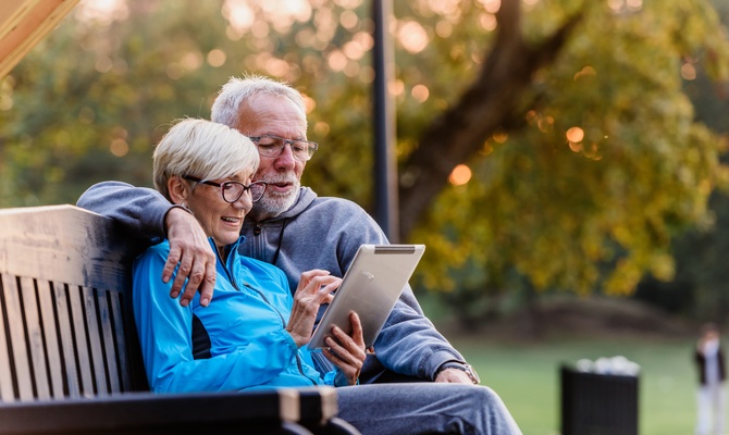An older man and woman sit together on a park bench and look at a tablet device. A tree stands behind them.
