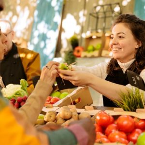 A woman smiles as she hands something to another woman across a stand full of potatoes and tomatoes.