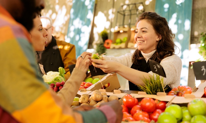 A woman smiles as she hands something to another woman across a stand full of potatoes and tomatoes.
