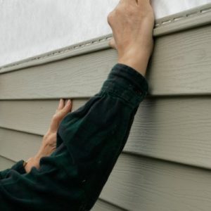 A close-up of two people's arms as they install siding onto a property. One person holds a hammer in their right hand.