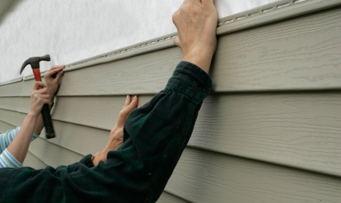 A close-up of two people's arms as they install siding onto a property. One person holds a hammer in their right hand.