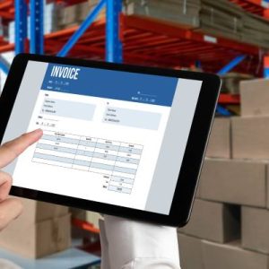 A close-up of a hand holding a tablet. On the screen is an invoice document. Boxes and pallets are in the background.