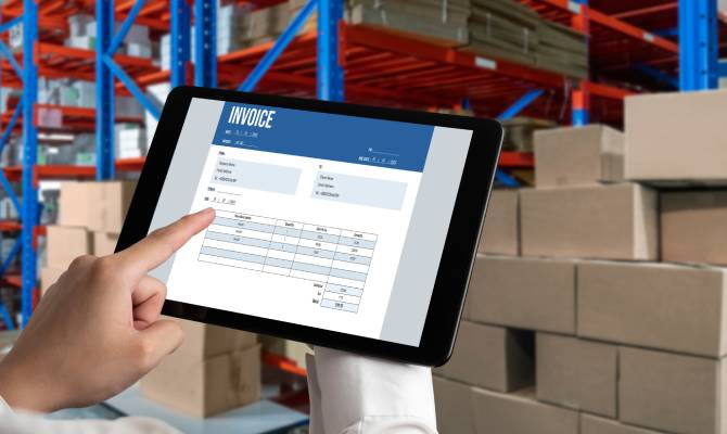 A close-up of a hand holding a tablet. On the screen is an invoice document. Boxes and pallets are in the background.