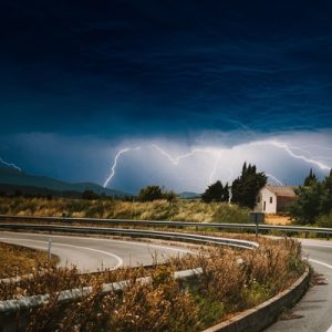 A white RV drives down a winding road. Dark blue clouds appear in the sky, with lightning strikes coming out of them.