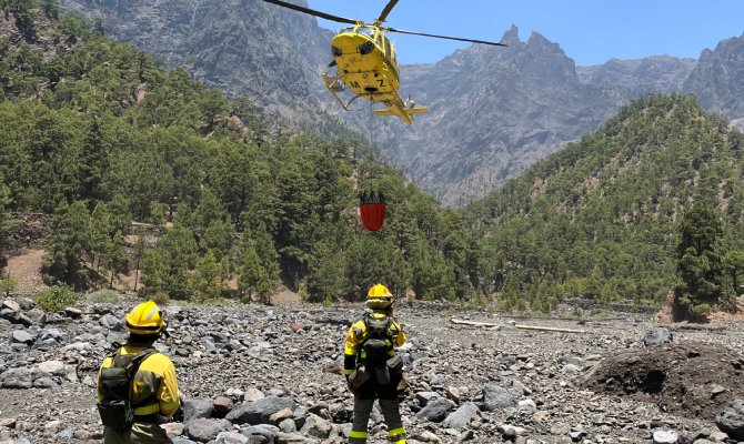 Two wildland firefighters wearing black and yellow protective equipment walk in mountains surrounded by forest.