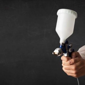 A person wearing a white protective gown holds an airbrush gun firmly in both hands and points it outward.