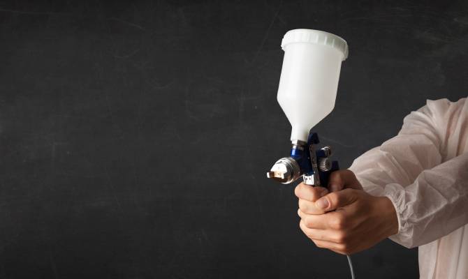 A person wearing a white protective gown holds an airbrush gun firmly in both hands and points it outward.