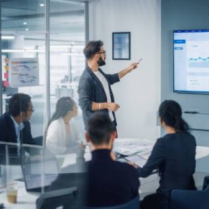 A man points with a pen at a line graph going up on the presentation screen during a business meeting.