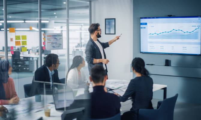 A man points with a pen at a line graph going up on the presentation screen during a business meeting.