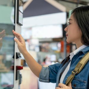 A young woman in a denim shirt stands in front of a smart vending machine, reaching out to use the touch screen.