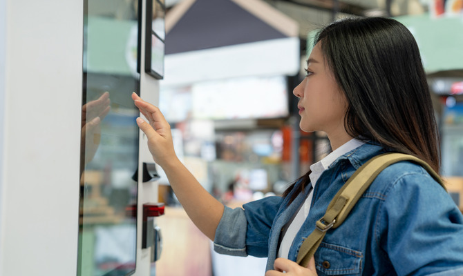 A young woman in a denim shirt stands in front of a smart vending machine, reaching out to use the touch screen.