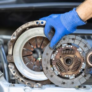 A close-up of a pair of hands with blue working gloves holding a used car clutch disc, pressure plate, and release bearing.