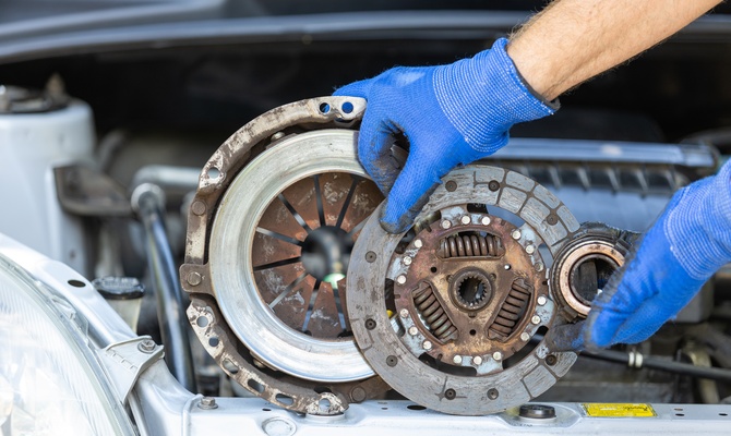 A close-up of a pair of hands with blue working gloves holding a used car clutch disc, pressure plate, and release bearing.
