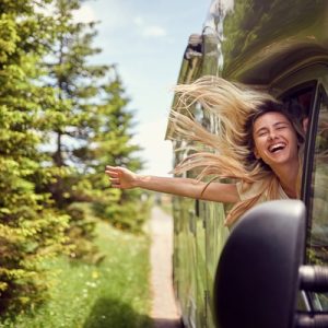 A smiling blonde-haired woman sticking her head and arm out the window of an RV as the wind blows through her hair.