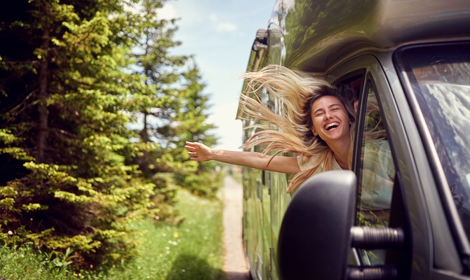 A smiling blonde-haired woman sticking her head and arm out the window of an RV as the wind blows through her hair.