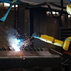 A yellow robotic arm welds a piece of metal with sparks flying off of it inside an industrial facility.