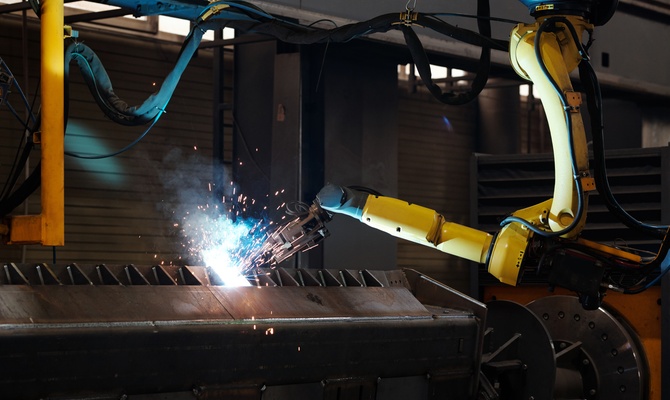 A yellow robotic arm welds a piece of metal with sparks flying off of it inside an industrial facility.