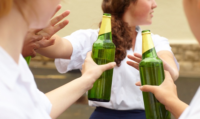 A teenage student turns down two bottles of alcohol being held out to her by two other people by holding up her hands.