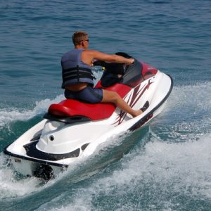 A man in a blue life jacket and blue swimsuit making waves while riding a white and red jet ski on a sunny day.