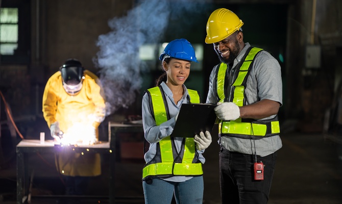 Two people wearing hard hats and hi-vis vests look at a clipboard. A welder stands behind them and welds something.