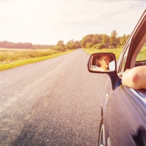 A young woman sits in the driver's seat of a blue sedan while driving down a long road with no other cars.