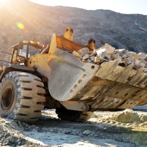 A close-up of a wheel loader machine hauling a large load of rocks over rough terrain while the sun shines above it.