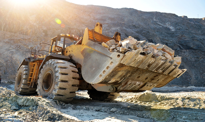 A close-up of a wheel loader machine hauling a large load of rocks over rough terrain while the sun shines above it.