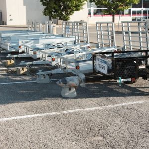 Several utility trailers lined up in a parking lot outside of a store. One trailer is black and the rest are silver.