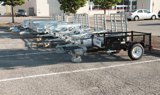 Several utility trailers lined up in a parking lot outside of a store. One trailer is black and the rest are silver.