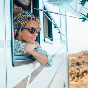 A happy young woman hanging her head outside of her parked RV window. She's wearing a bandana and sunglasses.