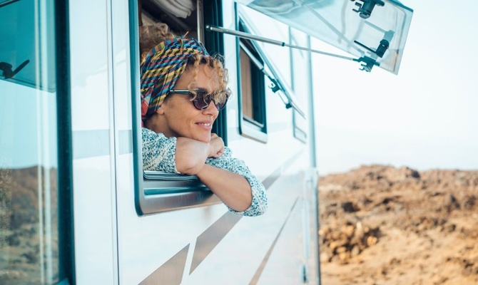 A happy young woman hanging her head outside of her parked RV window. She's wearing a bandana and sunglasses.