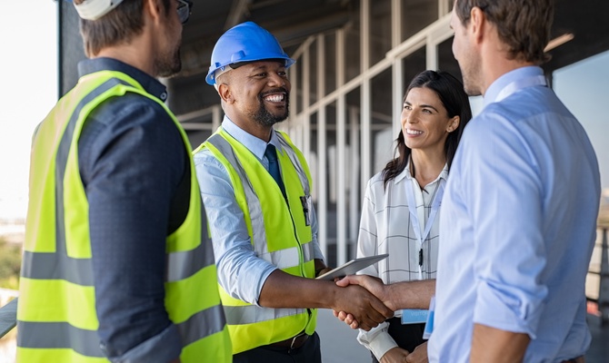 Contractors wearing safety vests and helmets on a construction site, with one giving a man a handshake next to a woman.