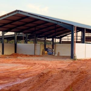 A pole barn with white siding and a peaked metal roof undergoing construction near a large dirt clearing.