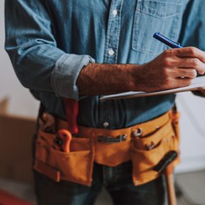 A close-up of a construction worker who is wearing a tool belt and denim shirt and writing on a clipboard.