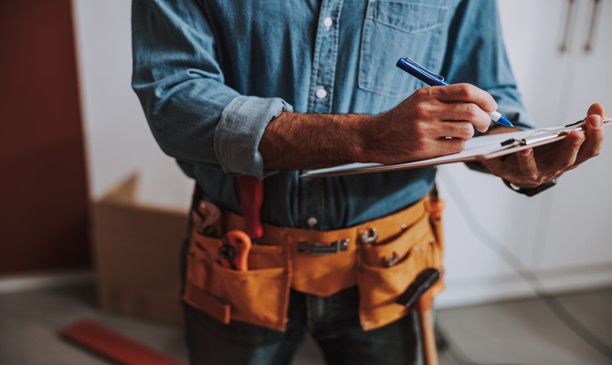A close-up of a construction worker who is wearing a tool belt and denim shirt and writing on a clipboard.