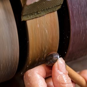 A lapidarist polishes a small tiger's eye stone on a grinding wheel, using water to lubricate the material.