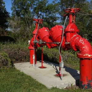 A large, red pipe sticks out above the ground surrounded by a cement block, tall grass, and mature trees.