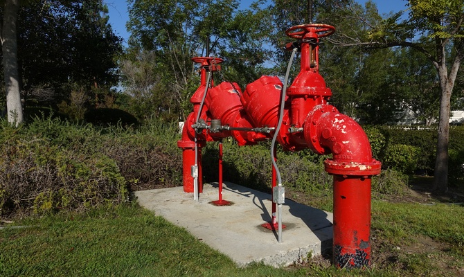 A large, red pipe sticks out above the ground surrounded by a cement block, tall grass, and mature trees.